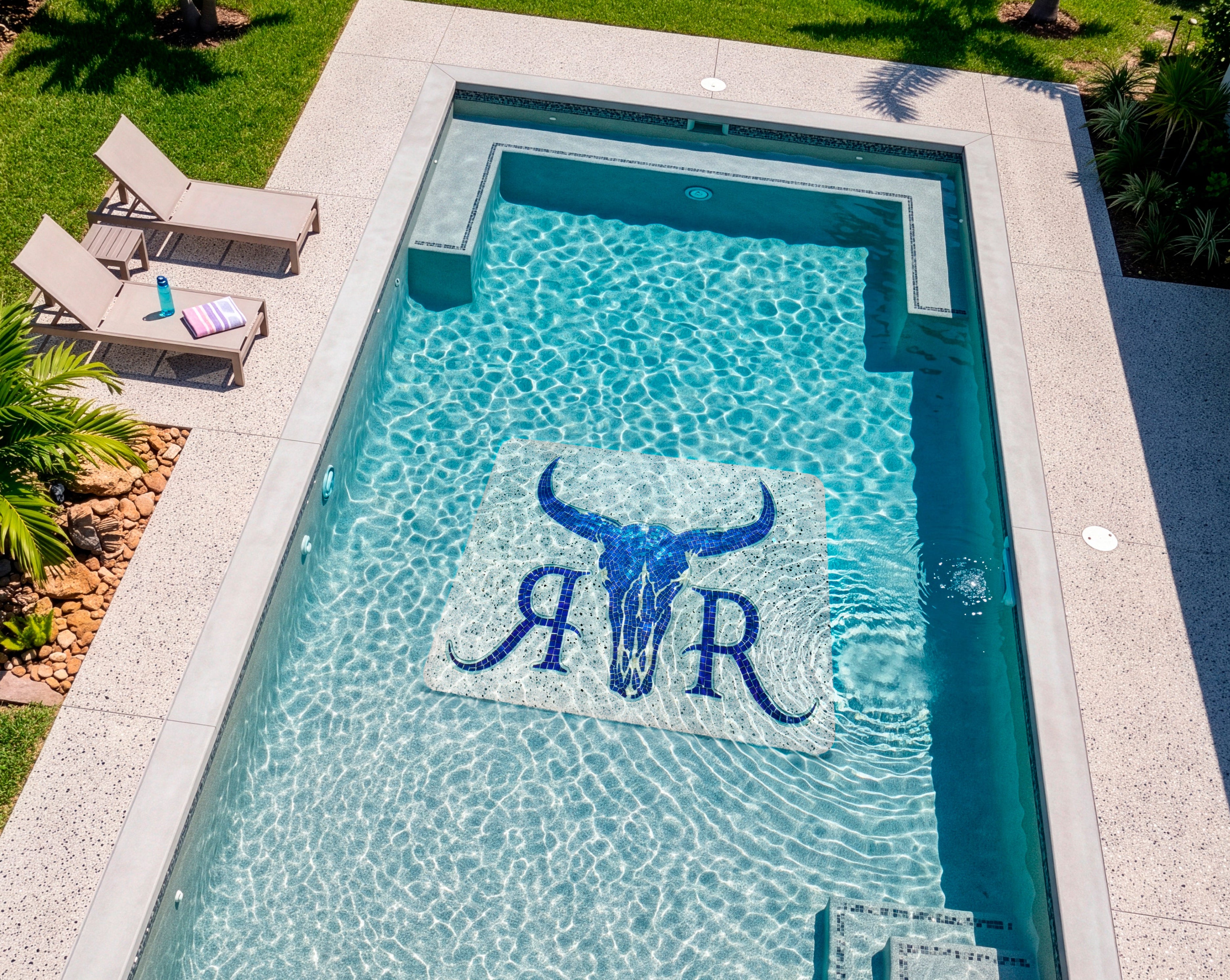 An aerial view of a rectangular outdoor swimming pool with clear turquoise water, a light stone deck, lounge chairs, grass, and a modern house. The pool floor features a large blue bull skull and the letters 'R&R' in blue.